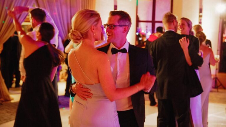 A couple dances joyfully at a wedding reception at Ritz-Carlton Bacara, surrounded by elegantly dressed guests enjoying the lively atmosphere created by a live wedding band. The vibrant lighting enhances the festive mood, showcasing the perfect setting for memorable moments. Ben Mallare Events & Entertainment specializes in providing top-notch live wedding bands, ensuring an unforgettable experience for couples and their guests.